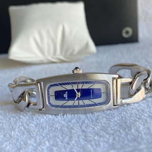May include: A silver-toned Breil watch with a rectangular blue and white face and a chain-link band. The watch face has the brand name and the word "ANTICHOC" printed on it. The watch is displayed on a white surface.