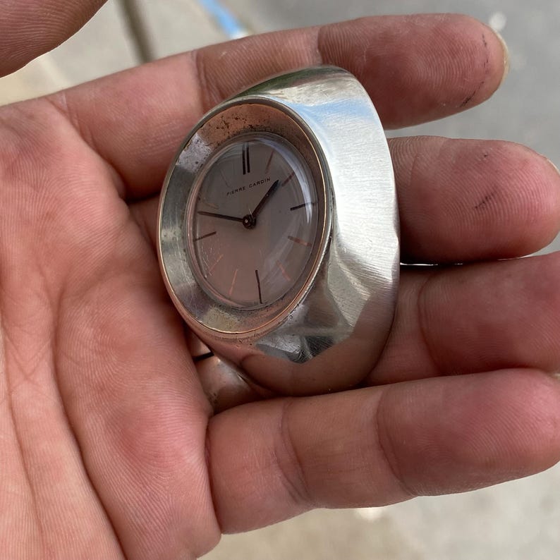 May include: A silver-toned ring watch with a light-colored face and black hands. The watch face is oval-shaped and features the brand name "Pierre Cardin". The ring has a wide, curved band.