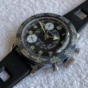 May include: A Cimier Sport chronograph wristwatch with a black dial, luminous hands, and Arabic numerals. The rotating bezel features a world time city list including London, Tokyo, and New York. It has a black leather strap with cutouts.