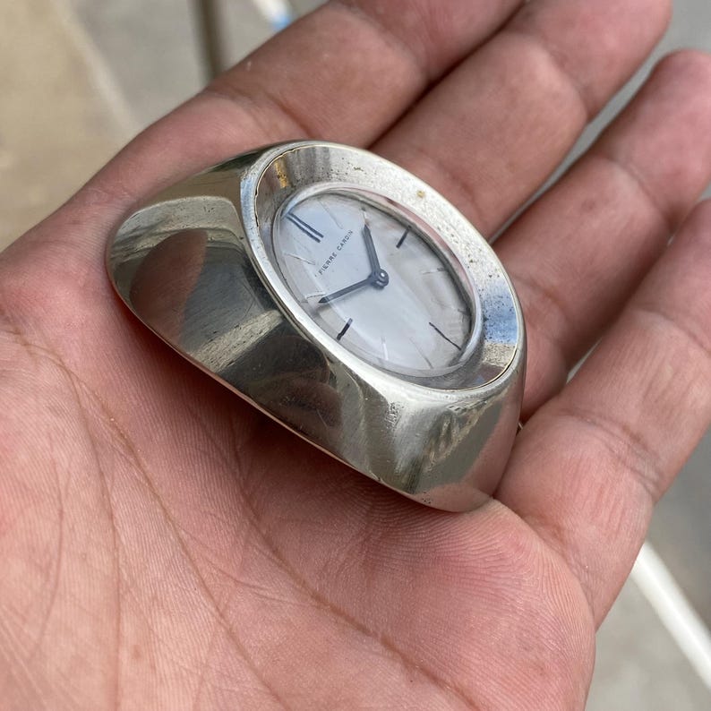 May include: A silver-toned Pierre Cardin ring watch with an oval face. The watch has a white dial with black hands and hour markers. The ring band is wide and rounded.