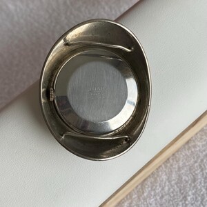 May include: A vintage silver-toned watch with a round face and an oval-shaped bezel. The watch face is silver with the text "JAEGER ITALY 33134". The watch is set against a white surface.