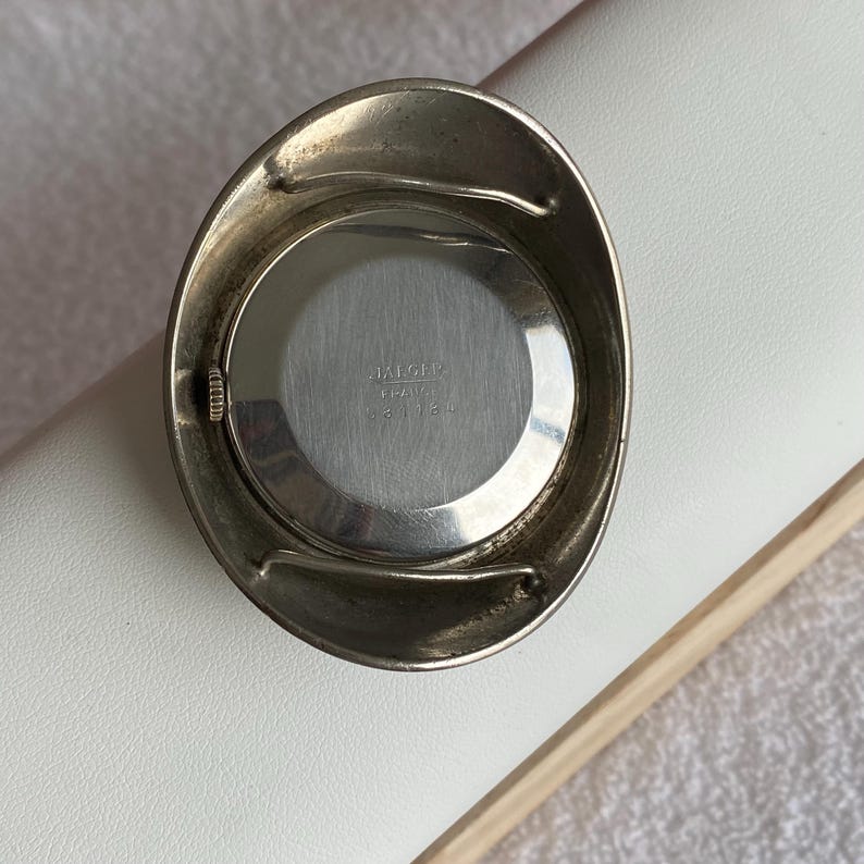 May include: A vintage silver-toned watch with a round face and an oval-shaped bezel. The watch face is silver with the text "JAEGER ITALY 33134". The watch is set against a white surface.