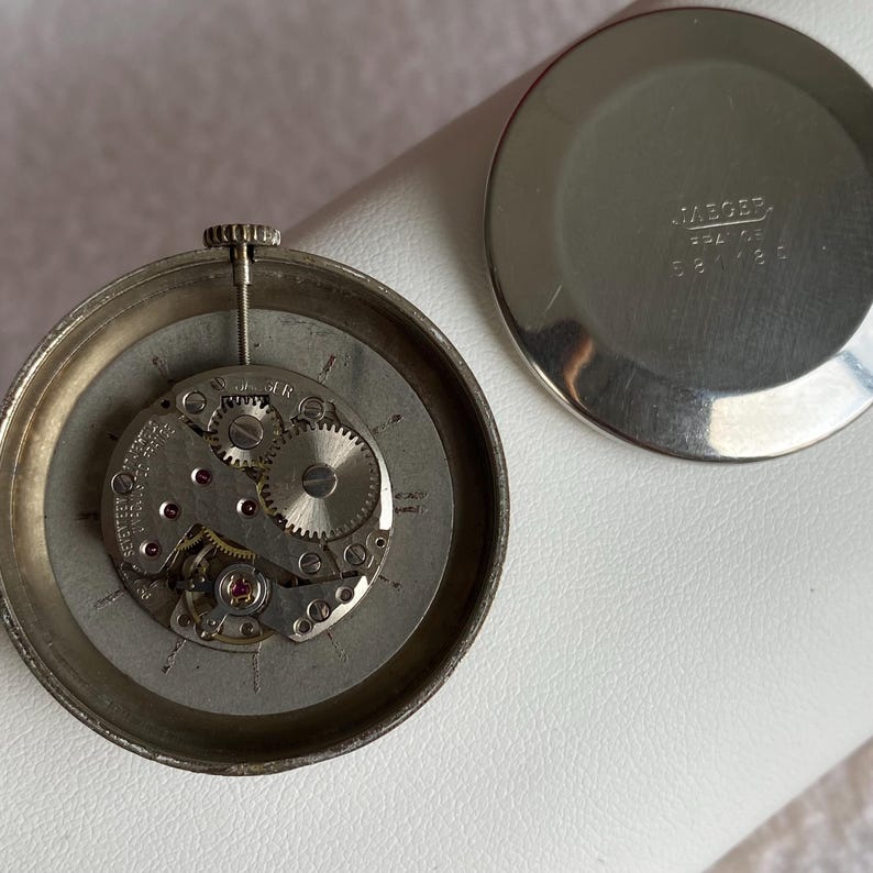 May include: An open vintage watch with its internal mechanism exposed. The silver-toned watch case surrounds the intricate gears, springs, and jewels of the movement. The watch back, also silver, is separate, with the name "JAEGER" and serial numbers visible.