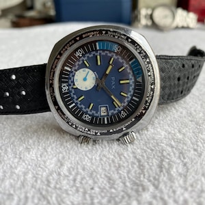 Puede incluir: Un reloj de pulsera PAX vintage con esfera azul, bisel de hora mundial y correa negra perforada. El reloj tiene una caja plateada y una ventana de fecha a las 6 en punto. La esfera del reloj muestra el texto "PAX".