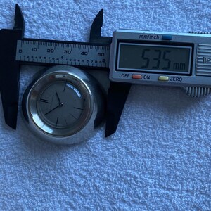 May include: A silver-toned wristwatch with a gray face is measured with a digital caliper. The caliper displays a measurement of 53.5 mm. The watch has black hands and a minimalist design.
