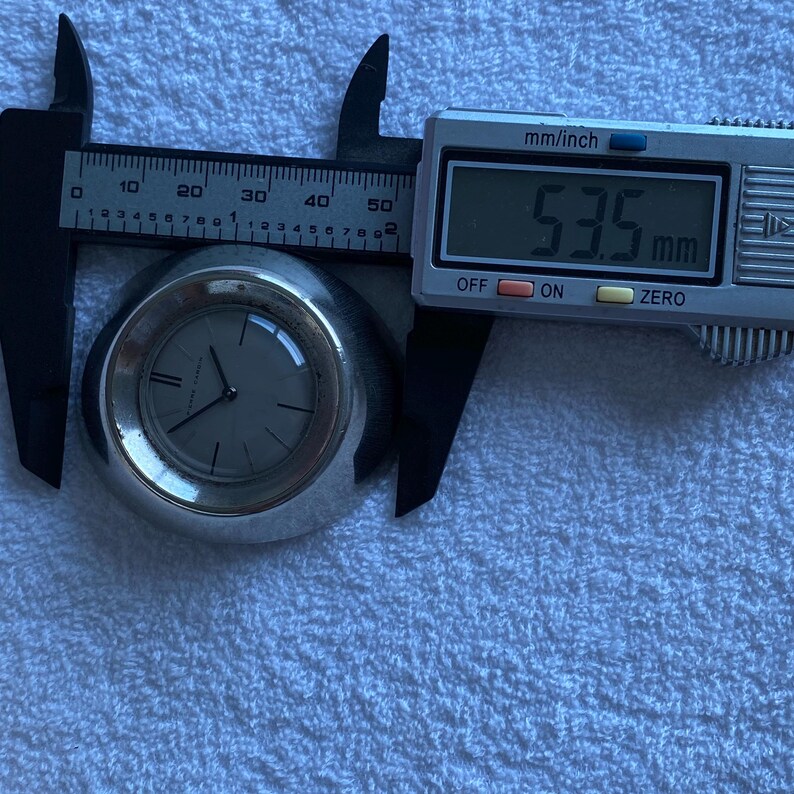 May include: A silver-toned wristwatch with a gray face is measured with a digital caliper. The caliper displays a measurement of 53.5 mm. The watch has black hands and a minimalist design.
