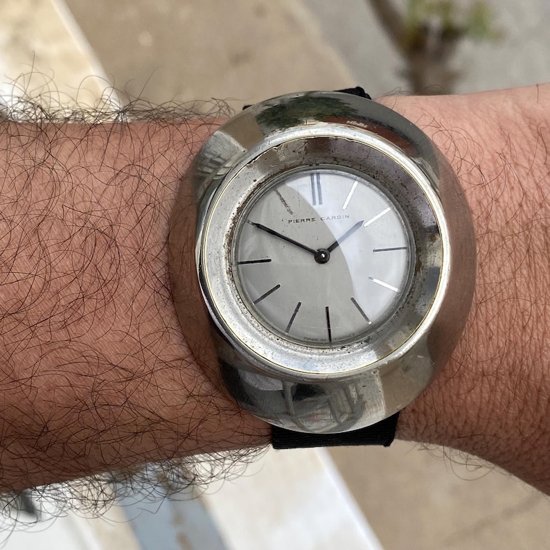 May include: A vintage Pierre Cardin wristwatch with a silver-toned case and a gray face. The watch has black hands and a black leather band. The watch is on a wrist.