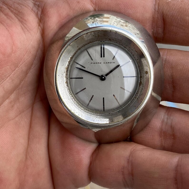 May include: A silver-toned Pierre Cardin wristwatch with a round, light-colored face. The watch features black hands and hour markers, with the brand name printed at the top. The watch has a unique, rounded design.