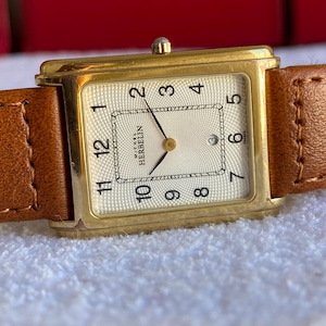 Könnte beinhalten: Eine rechteckige goldfarbene Armbanduhr mit strukturiertem Zifferblatt und braunem Lederarmband. Das Zifferblatt zeigt die Zahlen 1 bis 12, mit dem Markennamen "Michel Herbelin". Die Zeiger zeigen die Uhrzeit an.