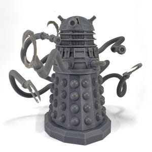 May include: A gray plastic model of a Dalek, a fictional robot from the British science fiction television series Doctor Who. The Dalek has a dome-shaped head with a single eye, a cylindrical body with a rotating base, and multiple tentacles.
