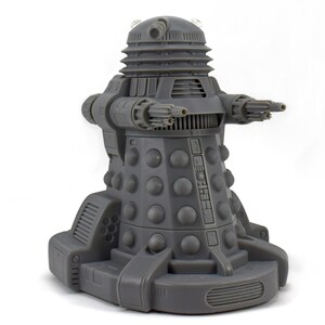 May include: A gray Dalek figurine, a fictional extraterrestrial race from the British science fiction television program Doctor Who. The figurine has a cylindrical body with a series of round bumps, a gun-like arm, and a manipulator arm.