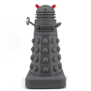 May include: A gray Dalek figurine with red antenna-like protrusions. The figurine has a cylindrical body with numerous gray spheres and is equipped with an arm and a weapon. The base is rectangular.