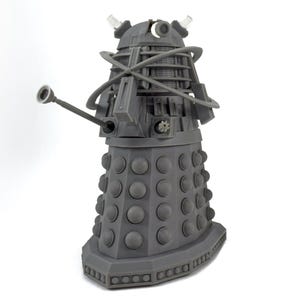 May include: A gray Dalek figurine, a fictional extraterrestrial race from the British science fiction television program Doctor Who. The figure has a cylindrical body with numerous round bumps, a gun-like arm, and a manipulator arm.