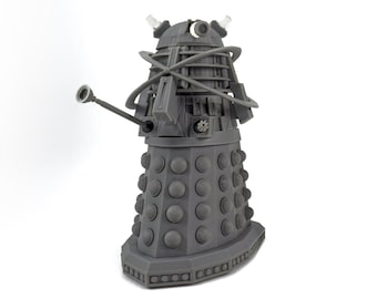 May include: A gray Dalek figurine, a fictional extraterrestrial race from the British science fiction television program Doctor Who. The figure has a cylindrical body with numerous round bumps, a gun-like arm, and a manipulator arm.