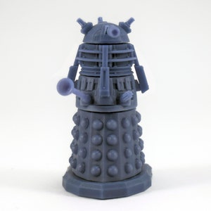 May include: A detailed, blue-grey figurine of a Dalek, a fictional extraterrestrial race from the Doctor Who series. The figure has a cylindrical base with numerous rounded protrusions, a central body section, and a head with various mechanical components.