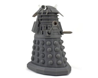 May include: A gray Dalek figurine, a fictional extraterrestrial race from the British science fiction television program Doctor Who. The figure has a cylindrical base with numerous round bumps, a central body, and a domed head with various attachments.