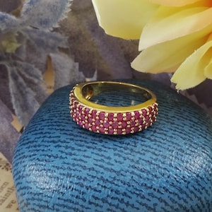 May include: A gold ring featuring three rows of small, bright red gemstones. The ring is set on a blue textured surface, with a yellow flower and a purple floral background.