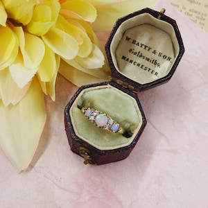 May include: A gold ring featuring three opal gemstones and small clear stones, presented in an open, antique, burgundy leather ring box. The box is lined with green velvet and has text that reads "W. BATTY & SON, Goldsmiths, MANCHESTER."