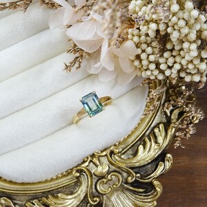May include: A gold ring featuring a rectangular emerald-cut blue-green gemstone set in a four-prong setting. The ring is positioned on a white velvet display surface, surrounded by dried cream-coloured flowers and a gold ornate frame.