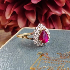 May include: A gold ring with a central, pear-cut, deep red gemstone, encircled by smaller, clear stones. The ring has a gold setting and a halo design. It rests on a blue surface with gold lettering, typical of vintage jewellery.