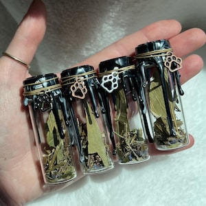 May include: Four small clear glass vials filled with dried herbs and leaves. Each vial is sealed with black wax that drips down the sides, wrapped with thin twine, and features a small metal paw print charm attached to the neck of the bottle.
