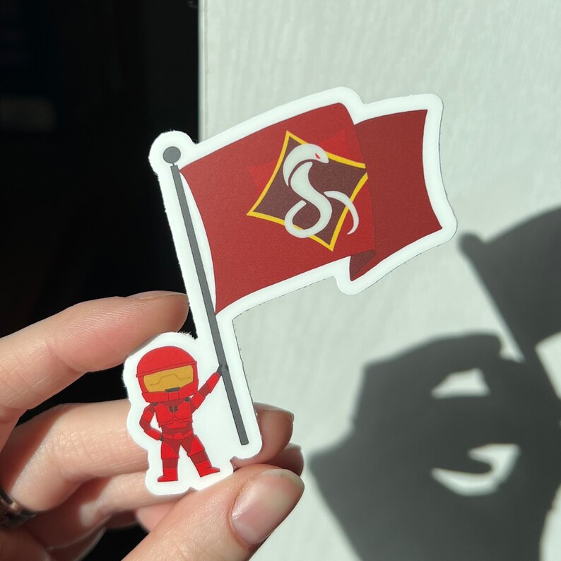 May include: A red and white sticker featuring a cartoon character holding a red flag with a white snake logo and the text "S" inside a diamond shape.
