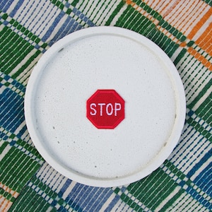 Puede incluir: Un posavasos de cerámica blanca con una señal de stop roja en el centro. La señal de stop tiene letras blancas que dicen "STOP".