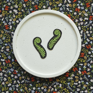 Może przedstawiać: Dwa zielone haftowane łaty z liśćmi paproci na białym ceramicznym talerzu. Talerz leży na czarnym tle z tkaniny z kwiatowym wzorem.