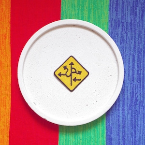 Puede incluir: Una señal de tráfico amarilla y negra con múltiples flechas apuntando en diferentes direcciones, sobre un fondo circular blanco. La señal está sobre una superficie rayada de colores.