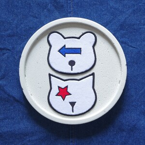 May include: White ceramic coaster with two felt patches. The top patch is a white bear with a blue arrow pointing left. The bottom patch is a white cat with a red star.