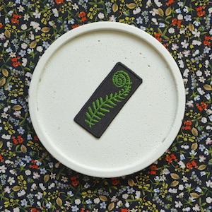 Może przedstawiać: Naszywka haftowana na czarno i zielono przedstawiająca paproć z zakręconą końcówką. Naszywka znajduje się na białym, okrągłym podłożu.