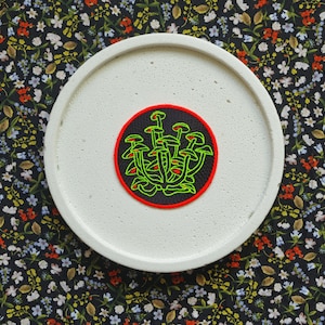 Peut inclure: Un dessous de verre en céramique blanche avec un patch brodé de champignons verts et rouges. Le patch est circulaire et a un fond noir.