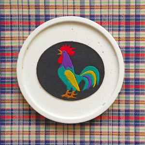 May include: A white ceramic coaster featuring an embroidered design of a colourful rooster on a black background. The rooster is green, yellow, purple and red.