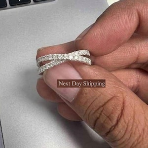 Puede incluir: Un anillo plateado con un dise&ntilde;o entrecruzado, incrustado con peque&ntilde;as piedras brillantes. El anillo se sostiene sobre un fondo gris, mostrando sus intrincados detalles y su estilo elegante.