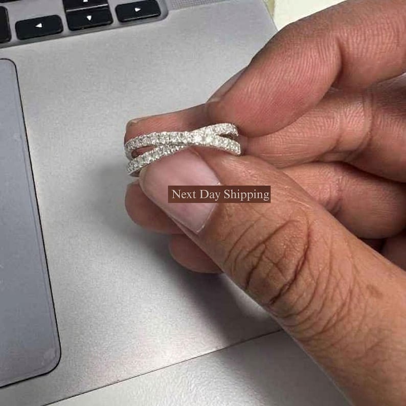 Puede incluir: Un anillo plateado con un dise&ntilde;o entrecruzado, incrustado con peque&ntilde;as piedras brillantes. El anillo se sostiene sobre un fondo gris, mostrando sus intrincados detalles y su estilo elegante.