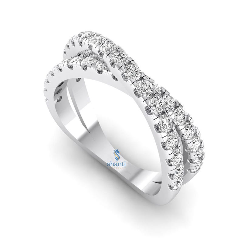 Puede incluir: Un anillo cruzado de oro blanco con dos bandas, cada una incrustada con peque&ntilde;as piedras preciosas redondas y transparentes. El anillo tiene una superficie pulida y reflectante y un dise&ntilde;o moderno. El anillo est&aacute; sobre un fondo blanco.