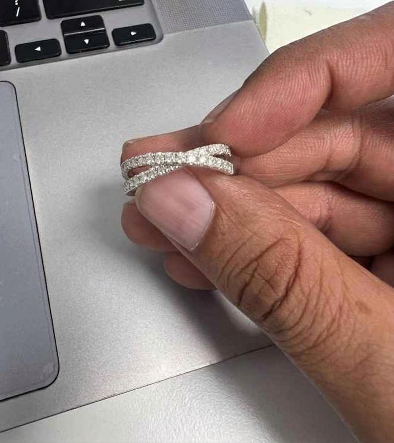 Puede incluir: Un anillo plateado con un dise&ntilde;o entrecruzado, incrustado con peque&ntilde;as piedras brillantes. El anillo se sostiene contra la superficie gris de un port&aacute;til, mostrando sus intrincados detalles y cualidades reflectantes. El anillo parece ser de metal y es una joya.