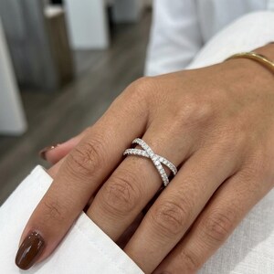 Puede incluir: Un anillo plateado entrecruzado con numerosas piedras peque&ntilde;as y transparentes. El anillo se lleva en un dedo y se ve una pulsera dorada. Las u&ntilde;as est&aacute;n pintadas de color marr&oacute;n oscuro. El fondo est&aacute; borroso.