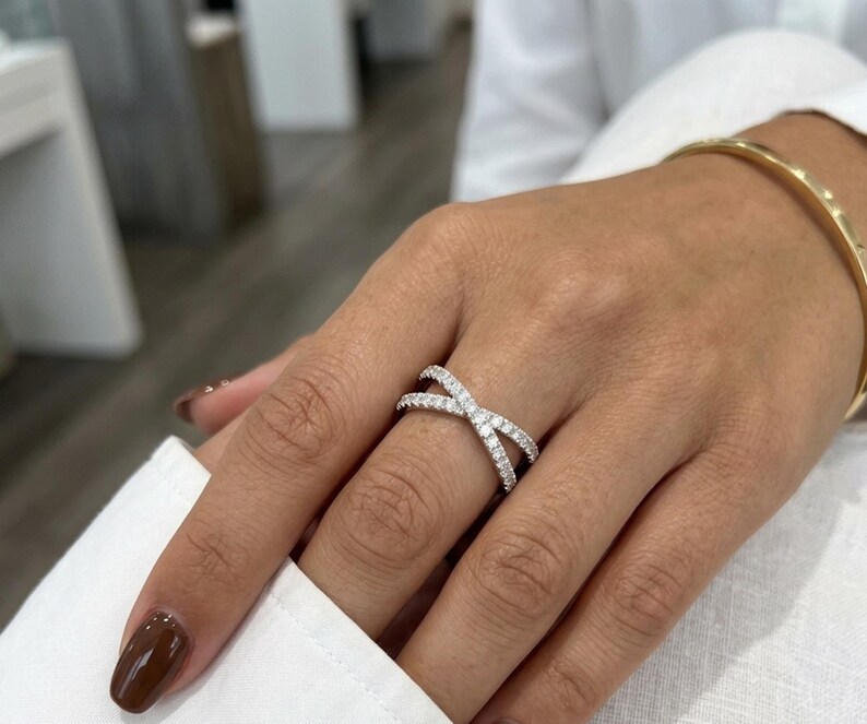 Puede incluir: Un anillo plateado entrecruzado con numerosas piedras peque&ntilde;as y transparentes. El anillo se lleva en un dedo y se ve una pulsera dorada. Las u&ntilde;as est&aacute;n pintadas de color marr&oacute;n oscuro. El fondo est&aacute; borroso.