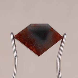 May include: A faceted, diamond-shaped gemstone with a mix of red and dark gray hues. The stone is held by a pair of silver metal tweezers, showcasing its polished surface and unique color patterns. The gemstone is likely for jewelry making.