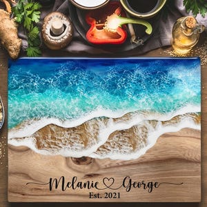 May include: A rectangular wooden cutting board with a resin ocean design. The top half features a blue and turquoise ocean with white waves, transitioning to a sandy beach design. The board is personalized with the names "Melanie & George" and "Est. 2021".