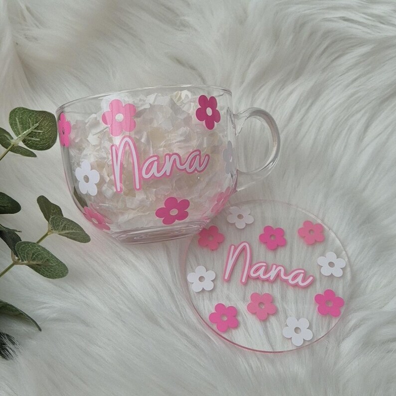 May include: A clear glass mug with a pink and white floral design and the word "Nana" in pink lettering. The mug comes with a matching pink and white floral coaster with the word "Nana" in pink lettering.