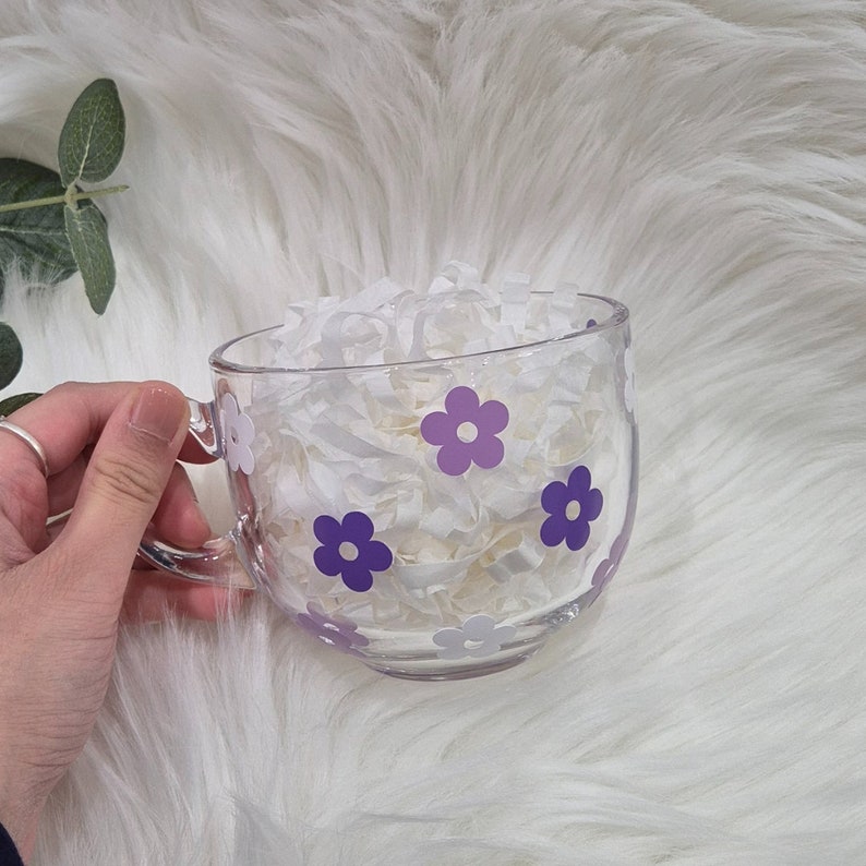 May include: A clear glass mug with a purple and white floral design. The mug is filled with white tissue paper.