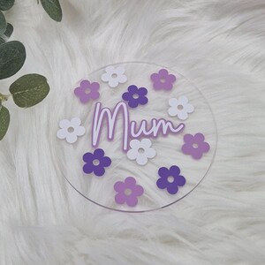 May include: A clear acrylic round coaster with the word "Mum" in white script and purple flowers around the edge.