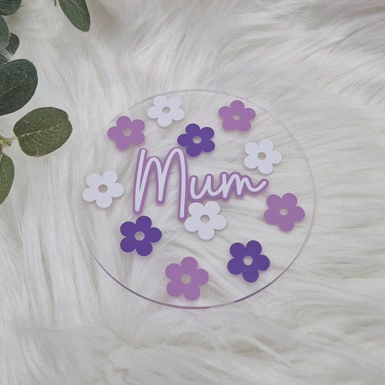 May include: A clear acrylic round coaster with the word "Mum" in white script and purple flowers around the edge.