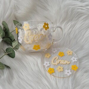May include: A clear glass mug with a white and yellow floral design and the word "Gran" written in white. The mug comes with a matching coaster.