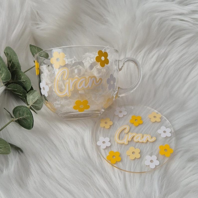 May include: A clear glass mug with a white and yellow floral design and the word "Gran" written in white. The mug comes with a matching coaster.