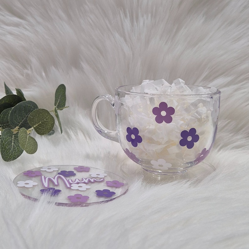 May include: A clear glass mug with a purple flower design and the word "Mum" on a matching coaster. The mug is filled with white tissue paper.