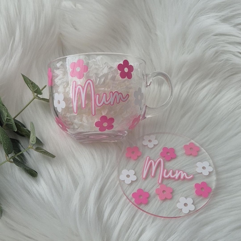 May include: A clear glass mug with a pink and white floral design and the word "Mum" in pink lettering. The mug comes with a matching coaster.
