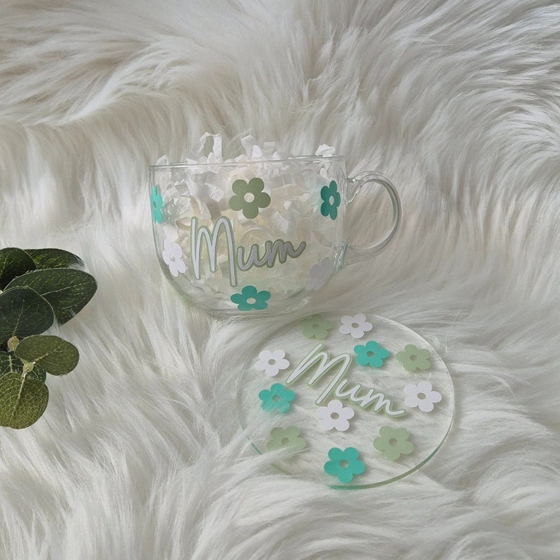 May include: A clear glass mug with a white and green floral design and the word "Mum" on the side. The mug is sitting on a white and green floral coaster.